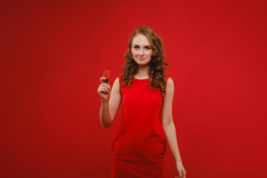 A beautiful girl in a red dress on a red background holds a strawberry in her hands and smiles.