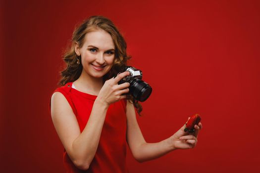 a smiling young woman with wavy hair holds a strawberry and photographs it, holding a delicious fresh strawberry on a bright red background.