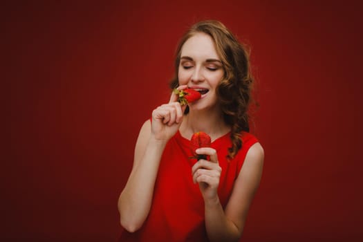 A beautiful girl in a red dress on a red background holds a strawberry in her hands and smiles.