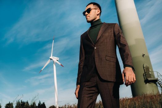 A man in a business suit with a green Golf shirt stands next to a windmill against the background of the field and the blue sky.Businessman near the windmills.Modern concept of the future