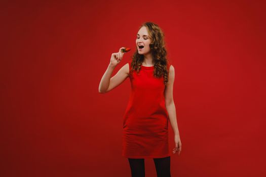 A beautiful girl in a red dress on a red background holds a strawberry in her hands and smiles.