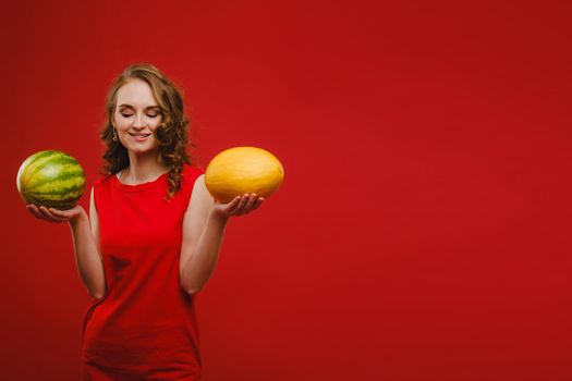 Photo of a cute girl in a red dress smiling with a watermelon and melon in her hands isolated on a bright red background