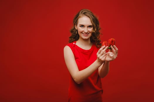 A beautiful girl in a red dress on a red background holds a strawberry in her hands and smiles.