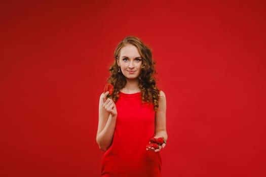 A beautiful girl in a red dress on a red background holds a strawberry in her hands and smiles.