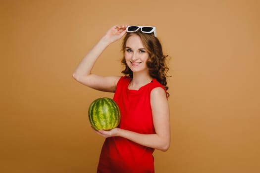 A beautiful girl in glasses and a red dress holds a watermelon in her hands.