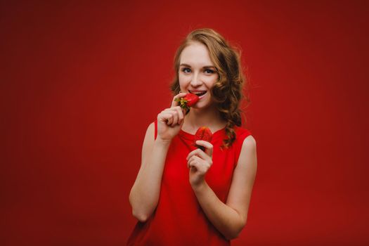 A beautiful girl in a red dress on a red background holds a strawberry in her hands and smiles.