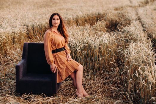 A girl in a orange dress is sitting on a chair in a wheat field in the evening.