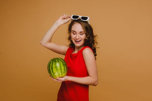 A beautiful girl in glasses and a red dress holds a watermelon in her hands.