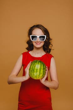 A beautiful girl in glasses and a red dress holds a watermelon in her hands.