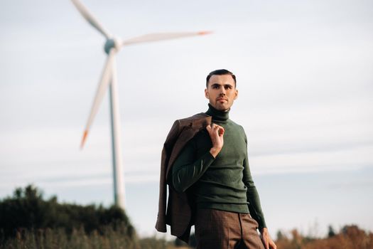 A man in a business suit with a green Golf shirt stands next to a windmill against the background of the field and the blue sky.Businessman near the windmills.Modern concept of the future