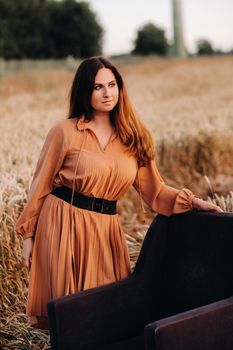 A girl in a orange dress stands next to a chair in a wheat field in the evening.