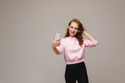 A smiling happy girl in a pink blouse takes a selfie on a smartphone on a gray background