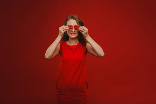 A beautiful girl in a red dress on a red background holds a strawberry in her hands and smiles.