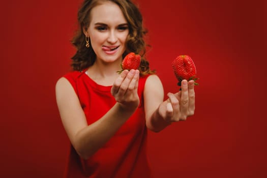 A beautiful girl in a red dress on a red background holds a strawberry in her hands and smiles.