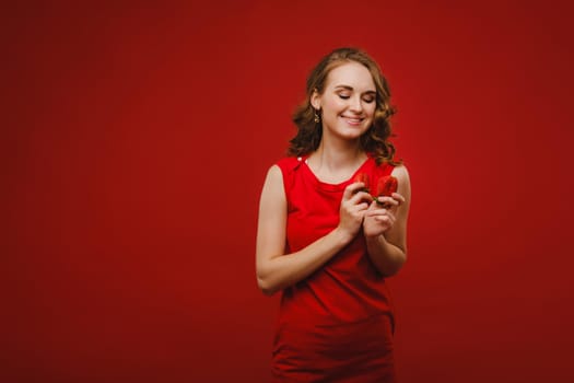 A beautiful girl in a red dress on a red background holds a strawberry in her hands and smiles.