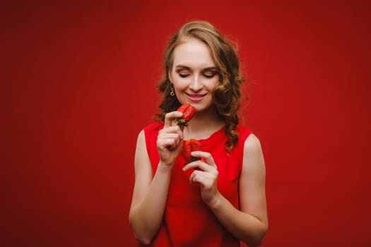 A beautiful girl in a red dress on a red background holds a strawberry in her hands and smiles.