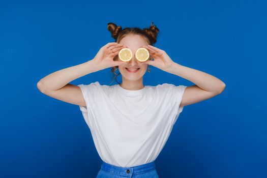 a young beautiful girl standing on a blue background holds lemons in her hands and covers her eyes with them.