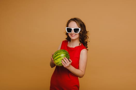 A beautiful girl in glasses and a red dress holds a watermelon in her hands.