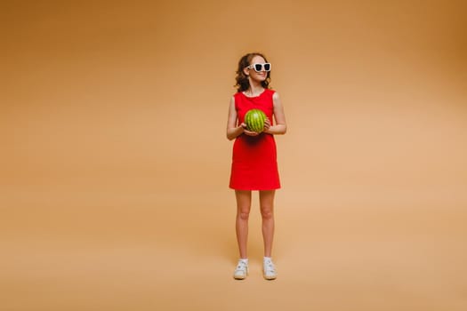 A beautiful girl in glasses and a red dress holds a watermelon in her hands.