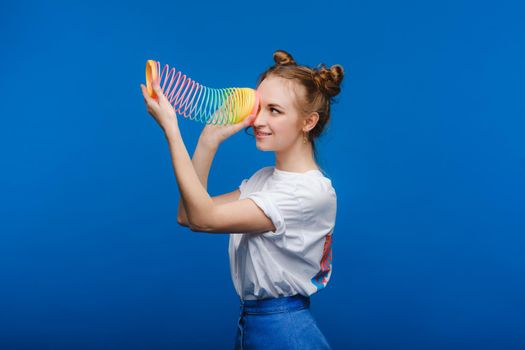 Beautiful young girl playing with a rainbow slinky, a toy of her childhood on a blue background.