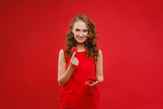 A beautiful girl in a red dress on a red background holds a strawberry in her hands and smiles.