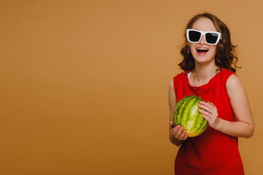 A beautiful girl in glasses and a red dress holds a watermelon in her hands.