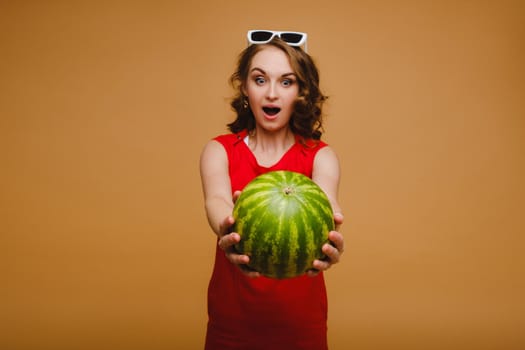 A beautiful girl in glasses and a red dress holds a watermelon in her hands.
