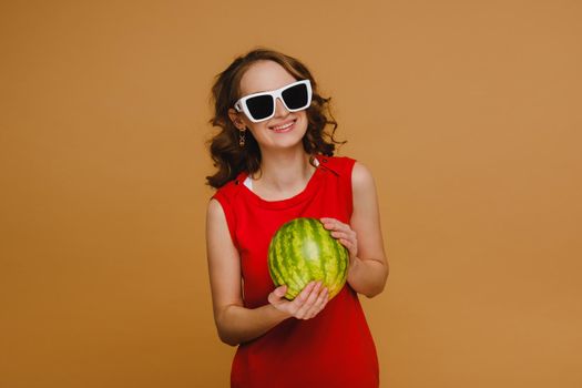 A beautiful girl in glasses and a red dress holds a watermelon in her hands.