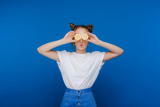 a young beautiful girl standing on a blue background holds lemons in her hands and covers her eyes with them.