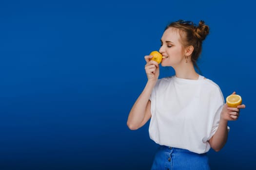 a young beautiful girl standing on a blue background holding lemons in her hand and biting