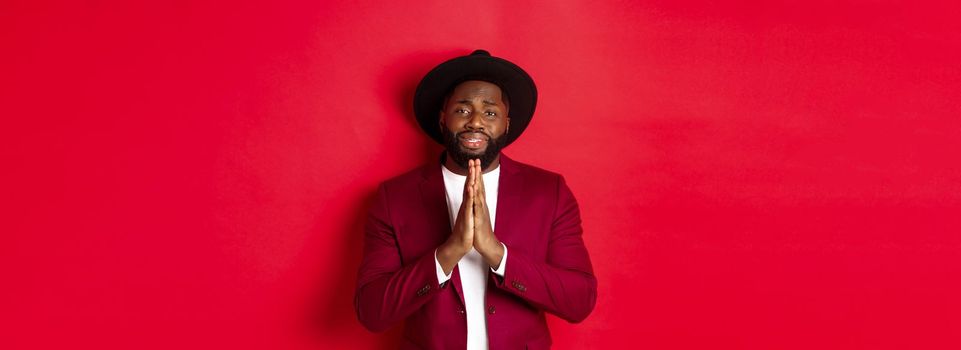 Miserable Black man asking for help, begging for favour and looking sad, standing against red background. Copy space