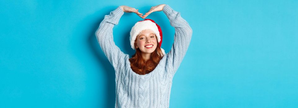 Winter holidays and Christmas Eve concept. Cute redhead teen girl in santa hat and sweater, making heart sign and smiling, wishing merry xmas, standing over blue background.