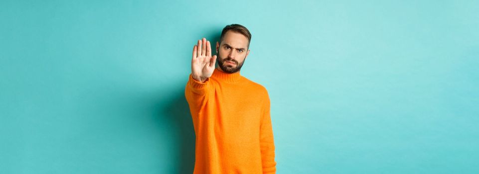 Serious frowning man showing stop gesture, telling no, prohibit and disagree, standing over light blue background.