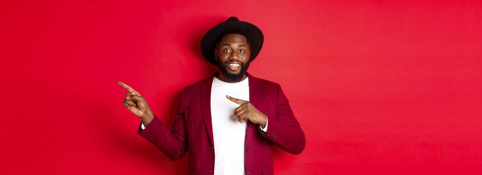 Winter holidays and shopping concept. Stylish african american man pointing fingers right at copy space for logo, standing over red background.