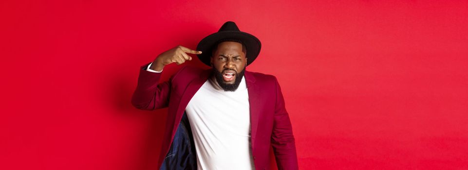 Angry Black man scolding person for being stupid, pointing at head and looking pissed-off, standing against red background.