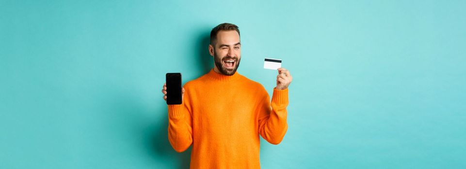 Online shopping. Handsome caucasian man showing credit card and smartphone, winking at camera, recommending something, standing over light blue background.