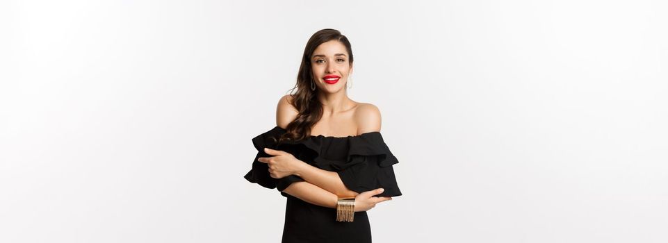 Beauty and fashion concept. Elegant and beautiful woman in black dress, makeup, hugghing herself and looking at camera with sensual gaze, standing over white background.
