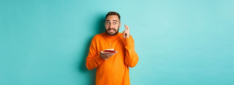 Handsome adult man celebrating birthday, blowing out candle on cake and making wish, standing against turquoise background.