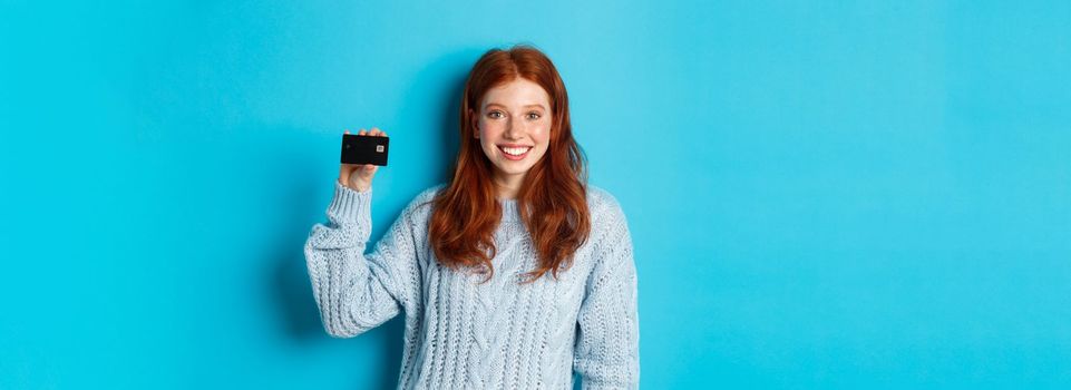 Cute redhead girl in sweater showing credit card, smiling at camera, standing over blue background.