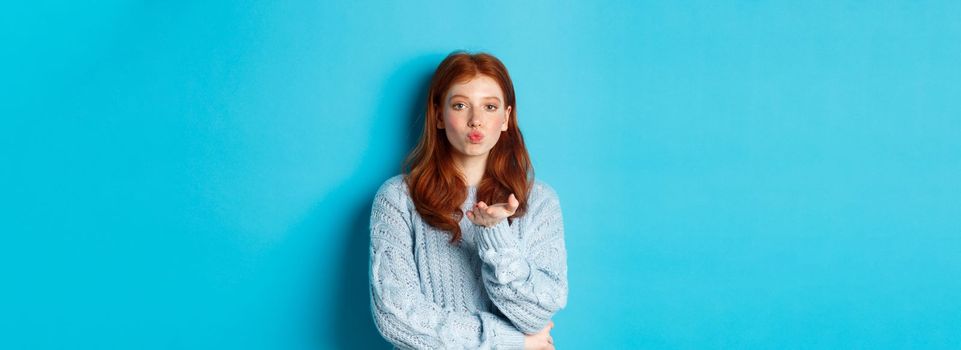 Lovely teen girl in sweater blowing air kiss, pucker lips and staring at camera, standing against blue background.