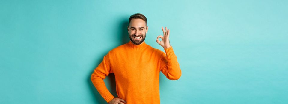 Image of confident handsome man showing ok sign, approve and agree, like something good, standing in orange sweater.