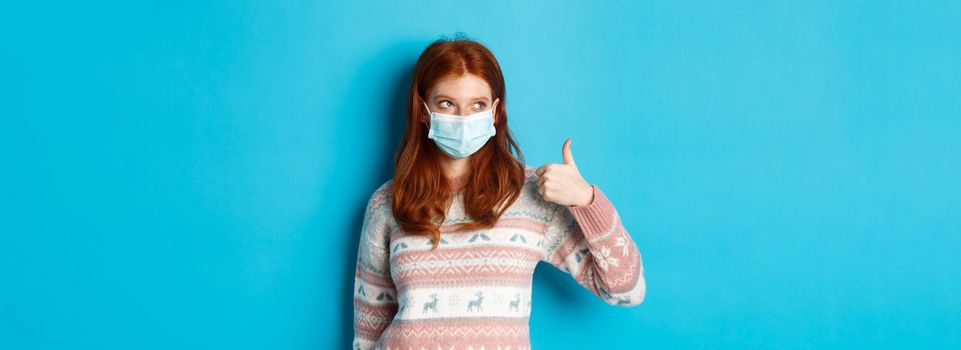 Winter, coronavirus and social distancing concept. Pretty redhead girl in face mask and sweater, looking at upper left corner promo and showing thumb up, praising good product.