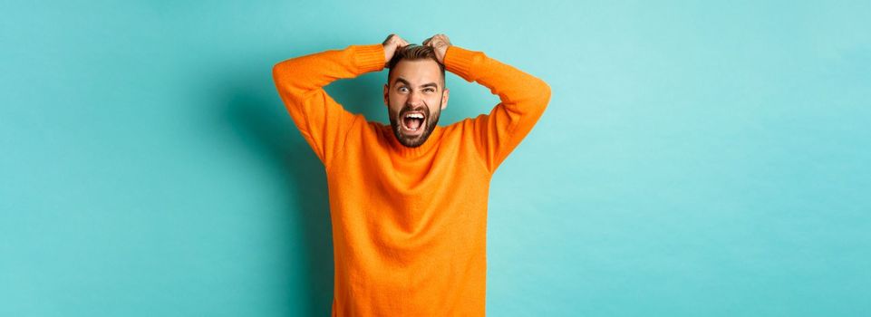 Frustrated man shouting, pulling out hair and screaming angry, losing temper and looking mad, standing over light blue background.