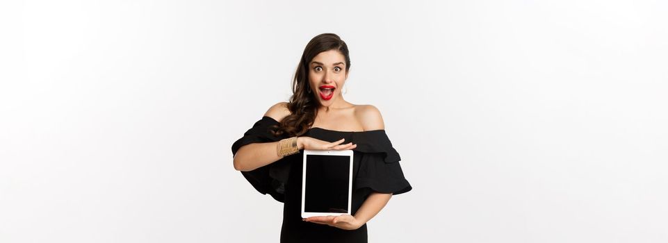 Fashion and shopping concept. Amazed young woman showing online website promo offer on tablet screen, looking excited camera, standing in black dress, white background.