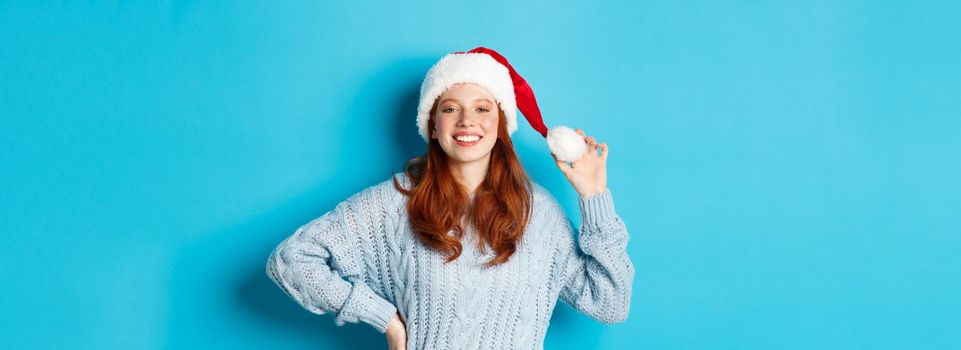 Winter holidays and Christmas Eve concept. Cheerful redhead teenage girl wearing Santa hat and smiling satisfied, standing in sweater against blue background.