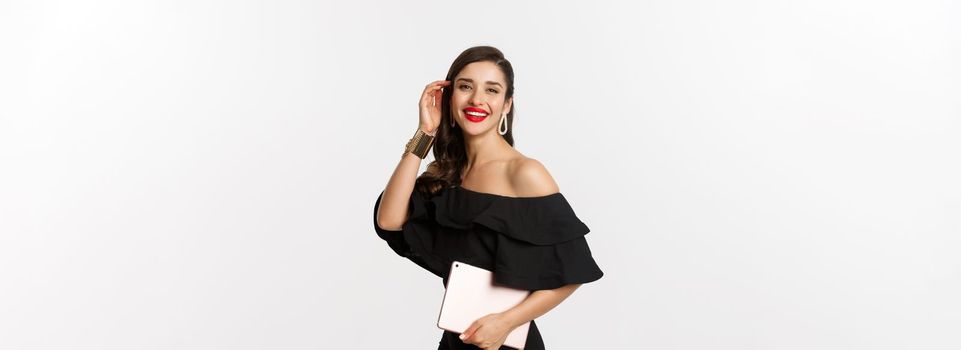 Fashion and shopping concept. Stylish young woman with glamour makeup, wearing black dress, holding digital tablet and smiling, white background.