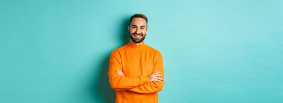 Handsome bearded male model in orange sweater, looking confident and smiling, cross arms on chest, standing over turquoise background.