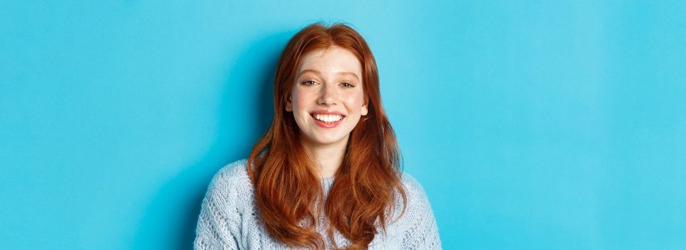 Close-up of cheerful teenage girl with red curly hair, smiling happy at camera, standing against blue background.