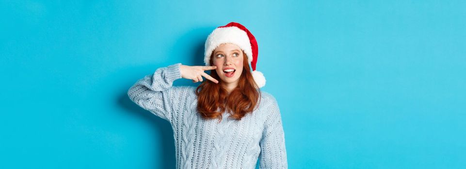 Winter holidays and Christmas Eve concept. Happy teenage girl with red hair, wearing santa hat, enjoying New Year, showing peace sign and gazing left at promo, standing over blue background.
