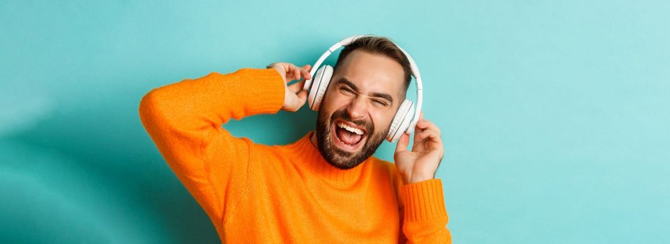 Close-up of handsome modern man listening music in headphones, standing in orange sweater over turquoise background.